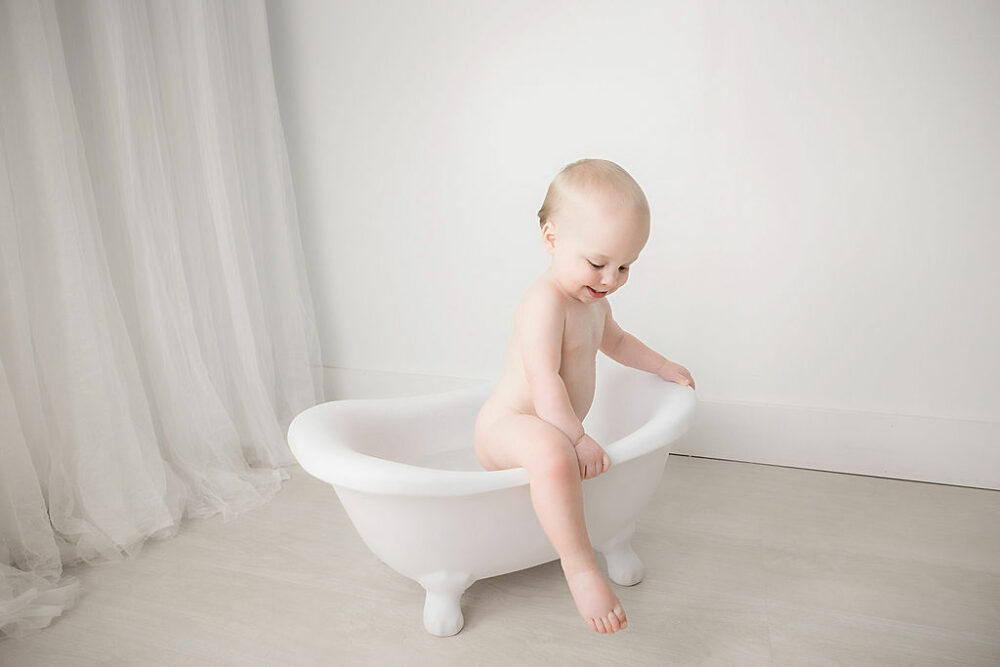 A baby in bath tub photography prop, smiling and playing for his minimalist first birthday session in Southampton, New Jersey.