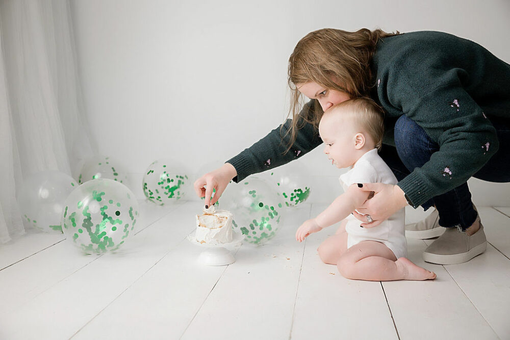 Side view of a mother and her son looking at his first birthday cake, they are both on the ground surrounded with balloons for their smash cake photography in Haddonfield, New Jersey.