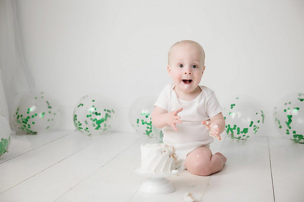 A toddler boy sitting on floor with a tiny birthday cake eating and having fun for his minimalist first birthday session in Bordentown, NJ.