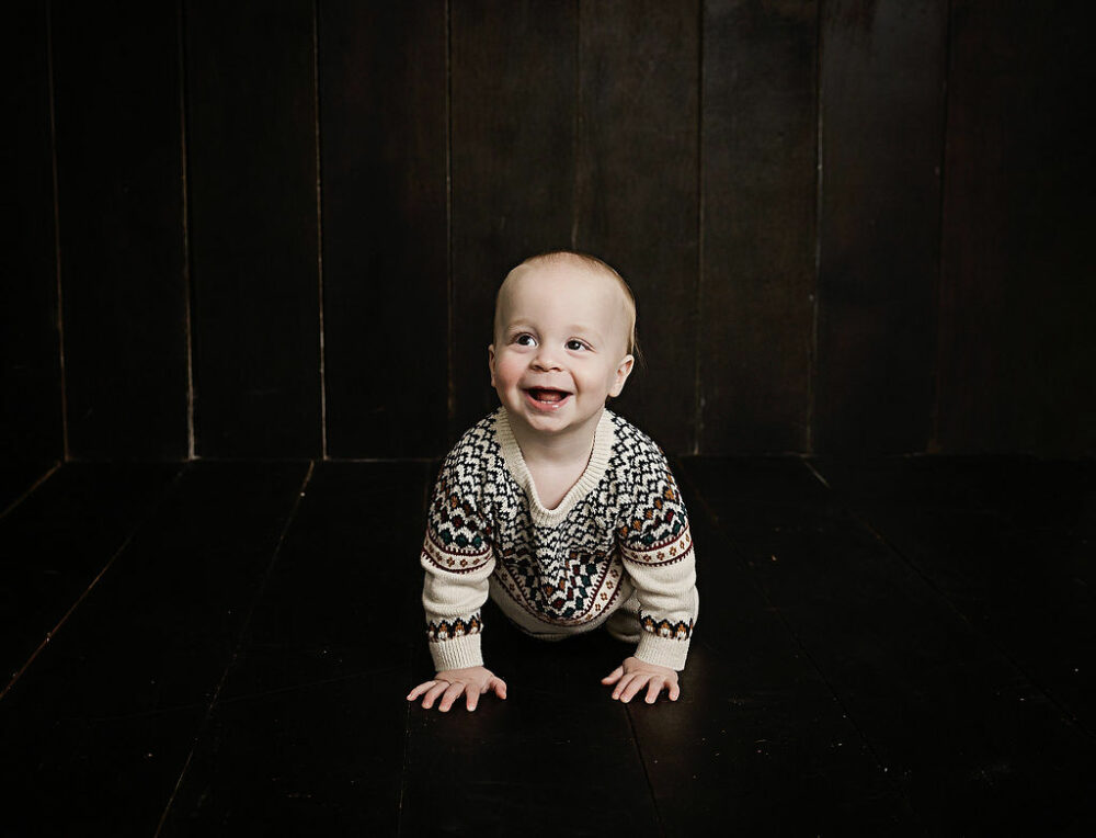 A lifestyle photo of a boy crawling on the floor and smiling wearing cute outfit for his in studio first birthday photo shoot in Cherry Hill, New Jersey.