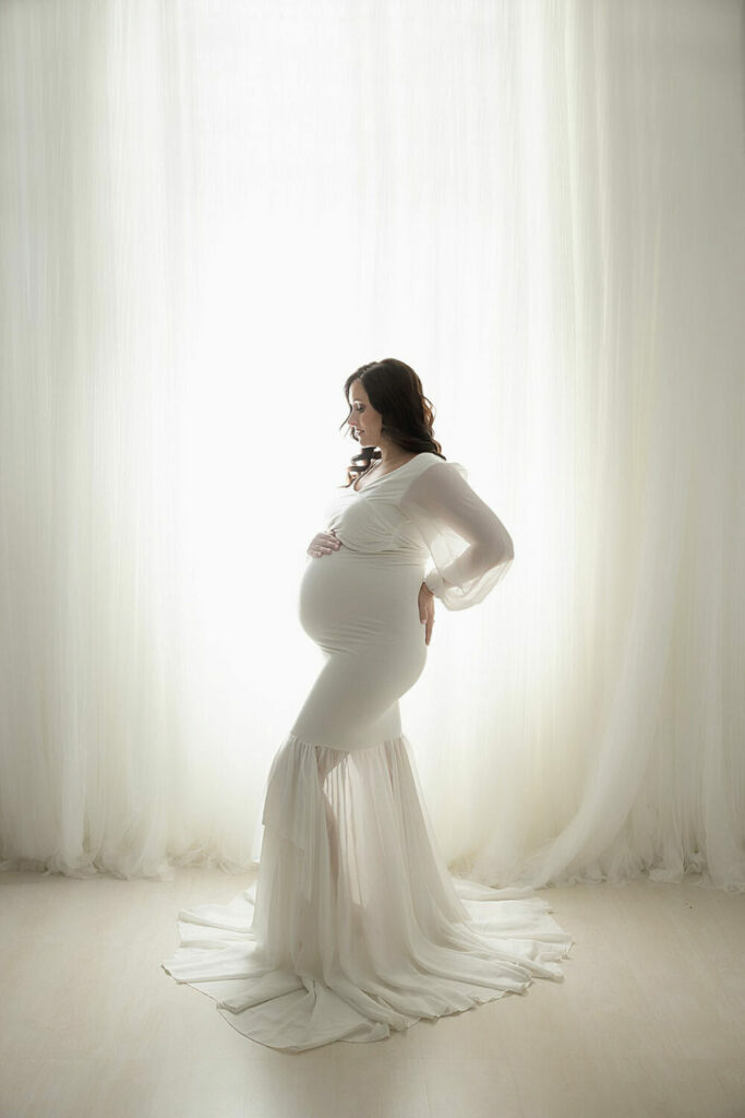 Hey side view of a woman doing pregnancy, poses wearing long gown against the light and bright background for her maternity session in Westampton, New Jersey.