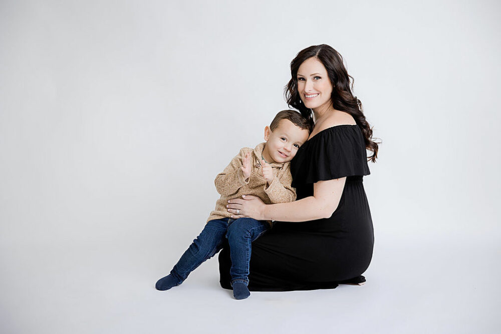A woman sitting on the ground with her toddler sign on her lap, both smiling for her maternity portraits taken in-studio in Mount Laurel, New Jersey.