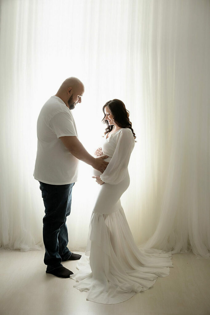 A man and woman facing each other, both looking down and smiling at her belly and holding it for their professional pregnancy pictures taken in Eastampton, New Jersey.