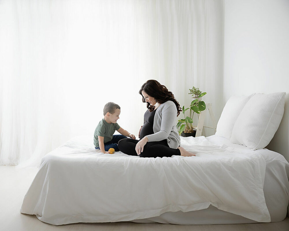 A woman and toddler boy sitting on the bed photography set against a light and bright background interacting for their baby bum photography session in Mount Holly, New Jersey.