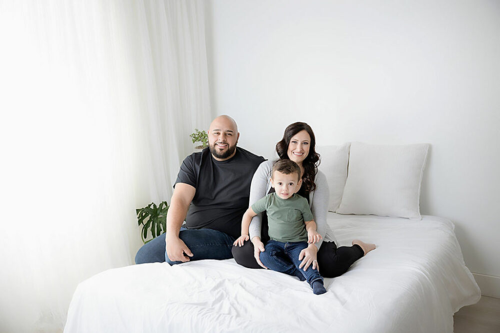 Men and women sitting on bed photography sat with their toddler son, smiling for their family pictures taken during her maternity photo shoot in Southampton, New Jersey.