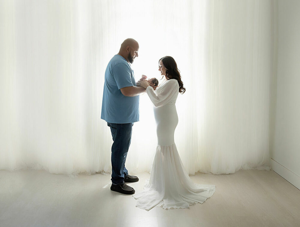 Side view of a man and a woman facing each other as they both hold their infant daughter, in their hands, posing for their newborn session in Haddonfield, New Jersey.