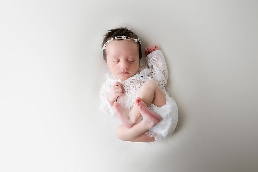 Infant girl sleeping on her back, wearing cute outfit and dainty headband with her feet curled up for her professional baby photography session in freehold, New Jersey.