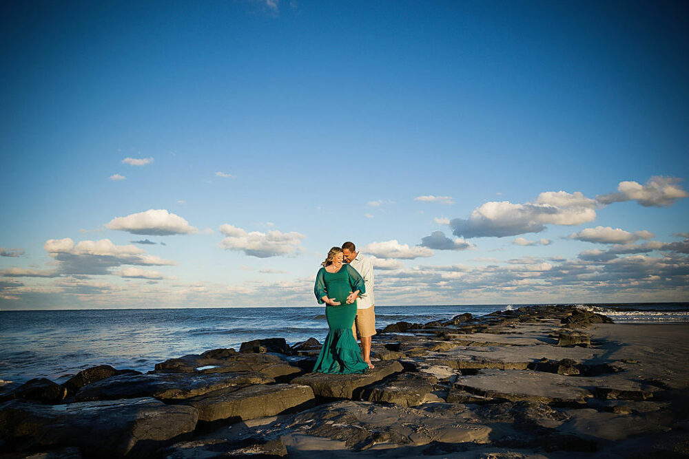 A man and a woman standing on rocks at the beach taking lifestyle portraits for her beach maternity session taken in Ocean City, New Jersey.
