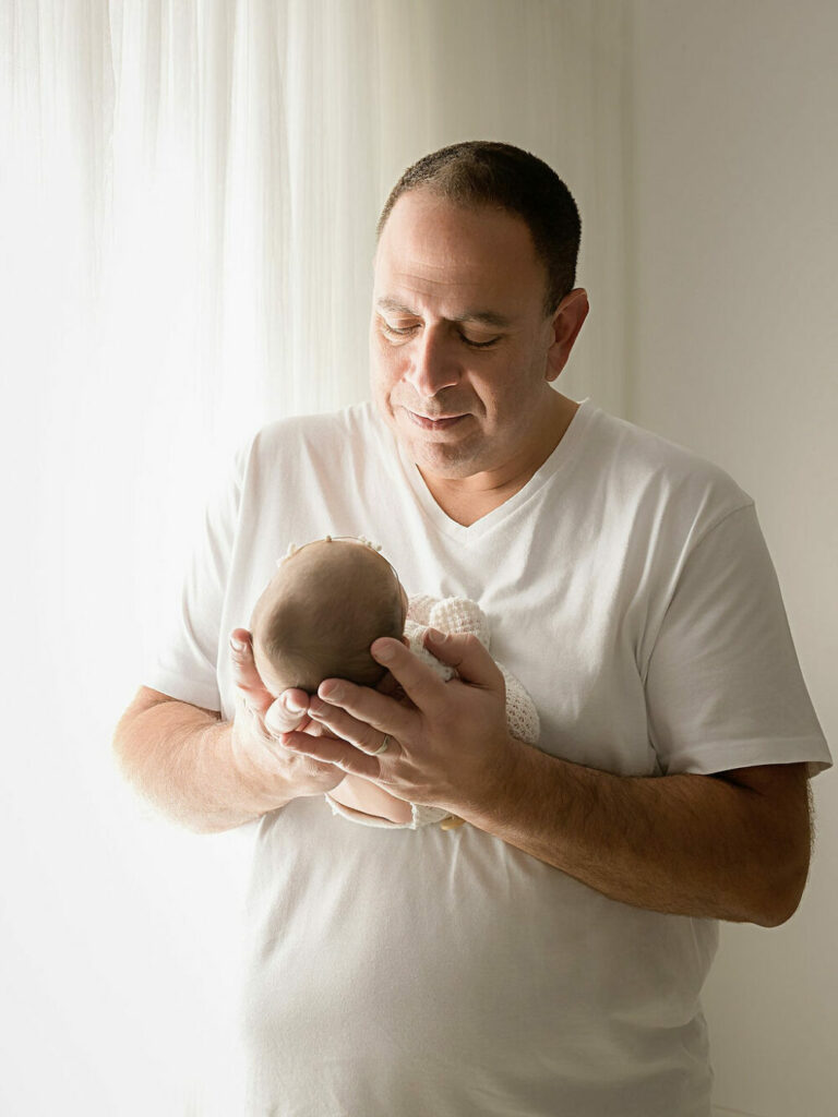 A father looking lovingly at his newborn daughter as he holds her in his hand for their rainbow baby newborn session taken in studio in Southampton, New Jersey.