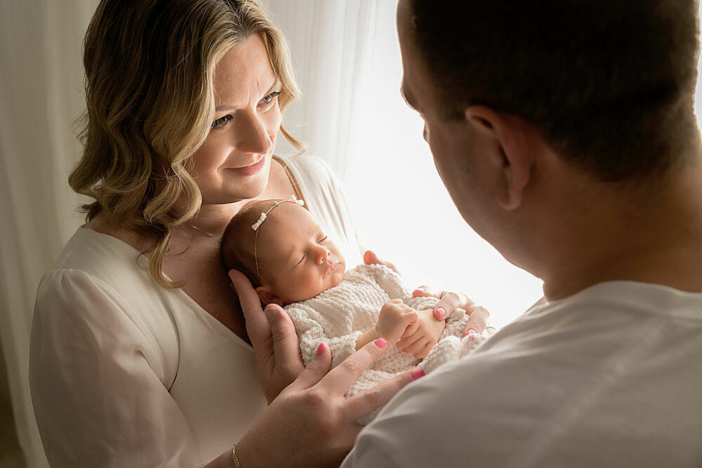 Close up of a family portrait of new parents holding their newborn daughter in their arms as the wife looks lovingly at her husband for their rainbow baby newborn session taken in Westampton, New Jersey.