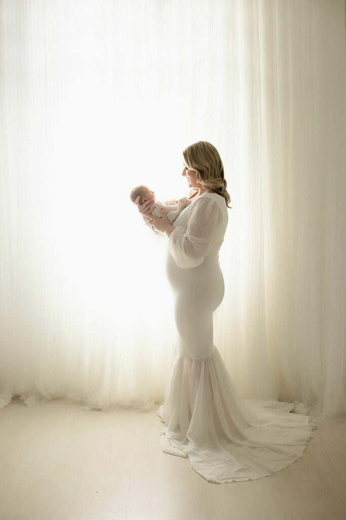A woman smiling and looking down at her newborn daughter wearing maternity dress against a light bright background before there famous portrait photographers in Eastampton, New Jersey.