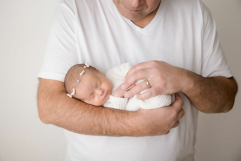A close-up of an infant sleeping in her father's arms for her rainbow baby newborn session taken in a professional studio in Burlington, New Jersey.