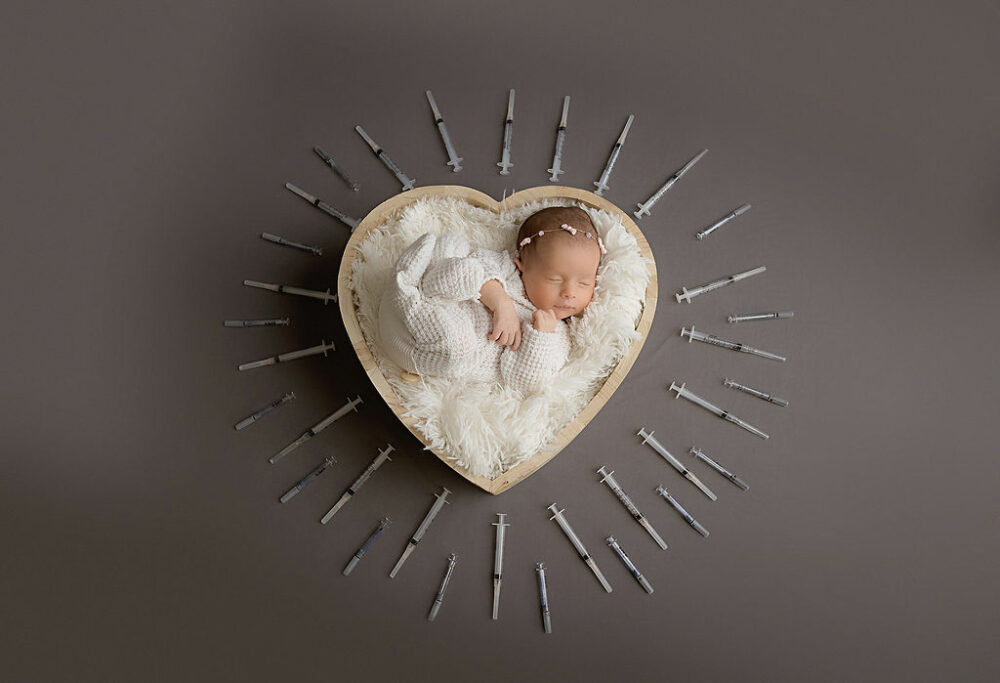 An infant girl sleeping in heart prop with IVF needles placed in a circle around her for her rainbow baby newborn session in Mount Holly, New Jersey.
