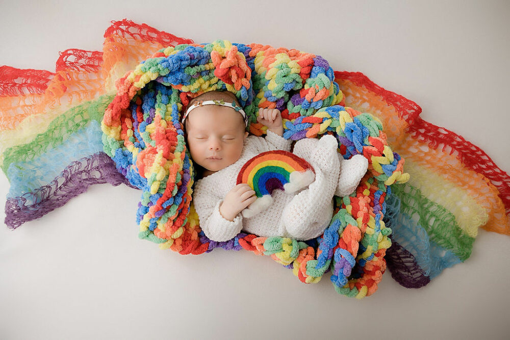 An infant sleeping on rainbow colored blankets and posing for her in-studio newborn session taken in Hamilton, New Jersey.