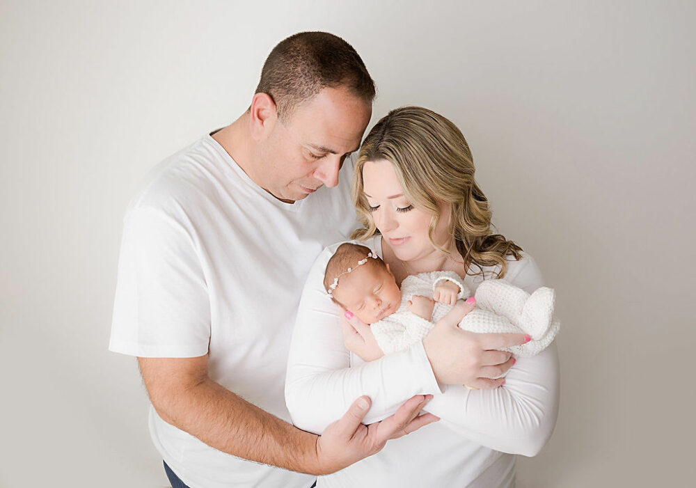 A classic family portrait of a mother and father looking down at their sleepy baby girl as she sleeps in her mothers arms for a rainbow baby newborn session in Morristown, New Jersey.