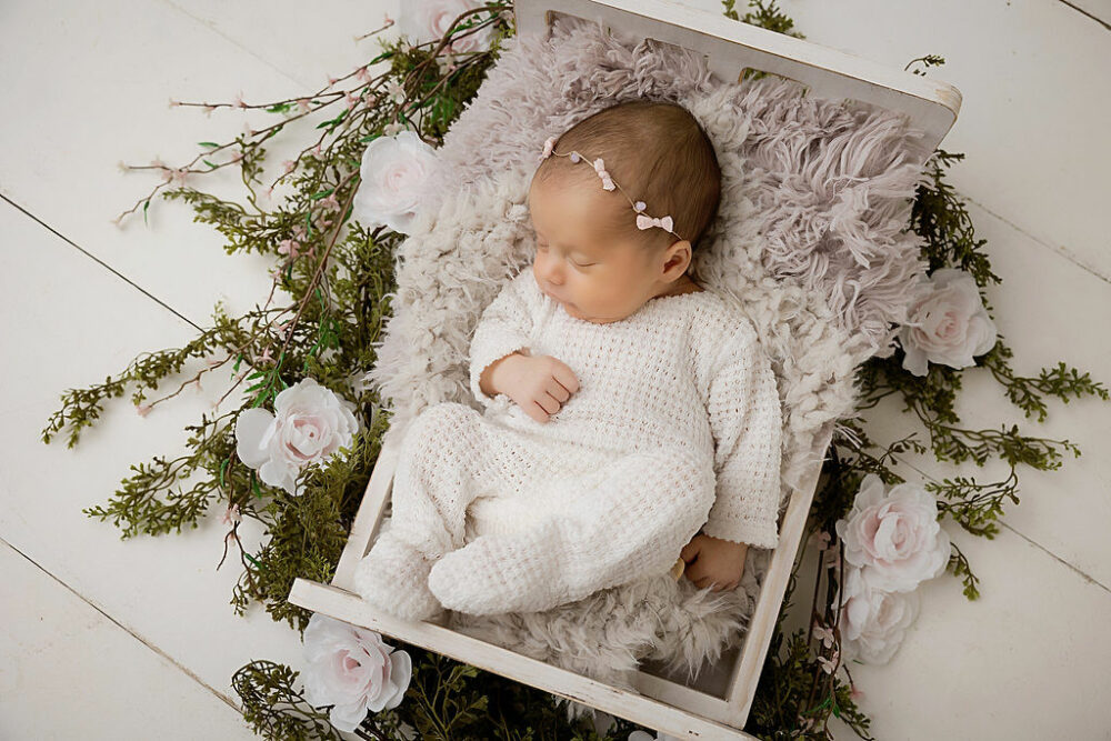 A close-up of an infant girl sleeping in a little crib adorned with flowers for her newborn photography session taken in-studio in Mount Laurel, New Jersey.