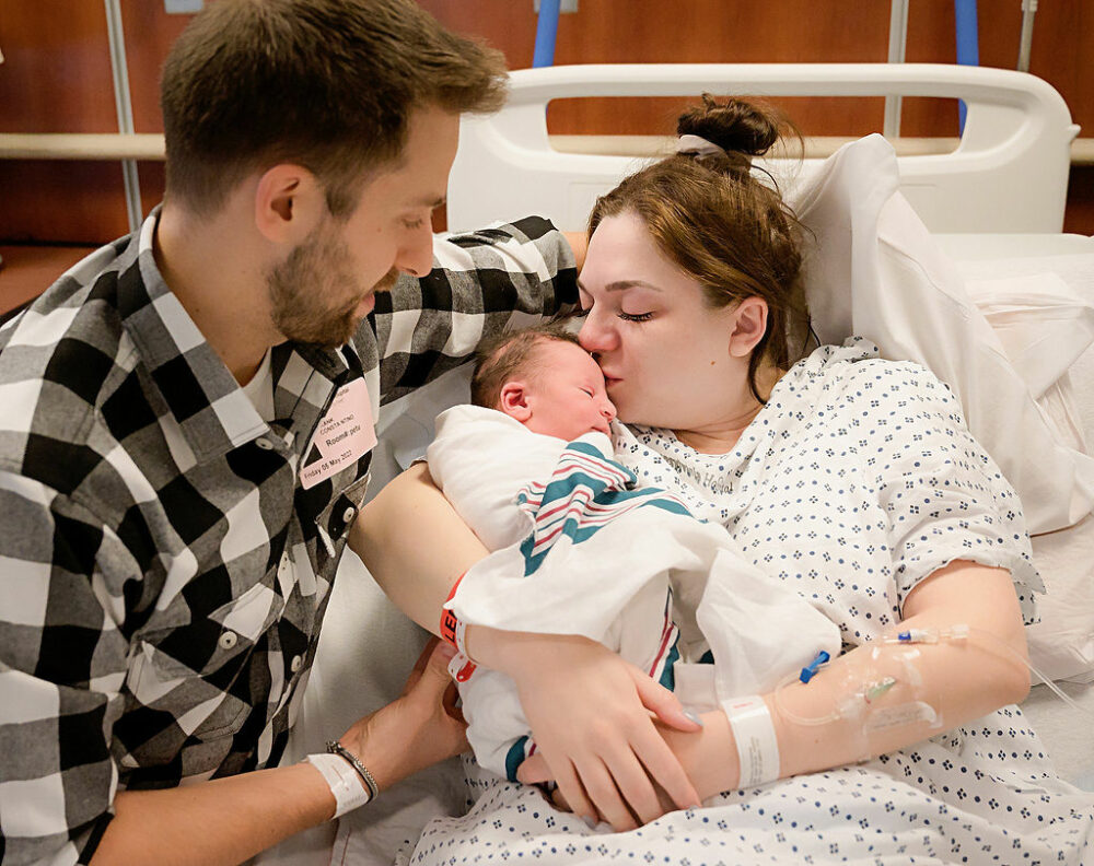 Family photo of a woman kissing her newborn son in hospital bed as her husband sits close for thier birth photography session in Mount Laurel, New Jersey.