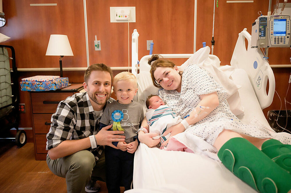 Family photography of a woman holding her newborn son in her hospital bed, while her toddler son, and husband stand next to them for their birth photography session in Deptford, New Jersey.