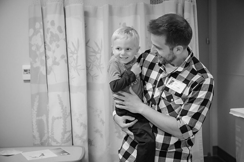 Black-and-white portrait of a father, carrying his toddler son as he meets his infant brother for the first time for their in hospital fresh 48 session in Waterford, New Jersey.
