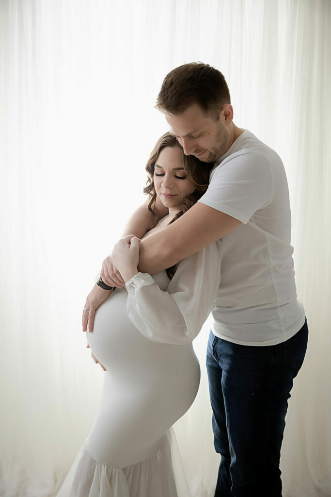 A pregnant woman wearing long white gown, while a man stand behind her as a both hold her belly for their professional maternity photos in Glocester, New Jersey.