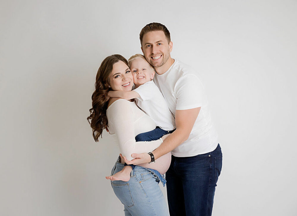 A man and a woman, hugging while holding their toddler in between them wearing jeans and matching T-shirts against a white backdrop for their black and white maternity session, taken in Southampton, New Jersey.