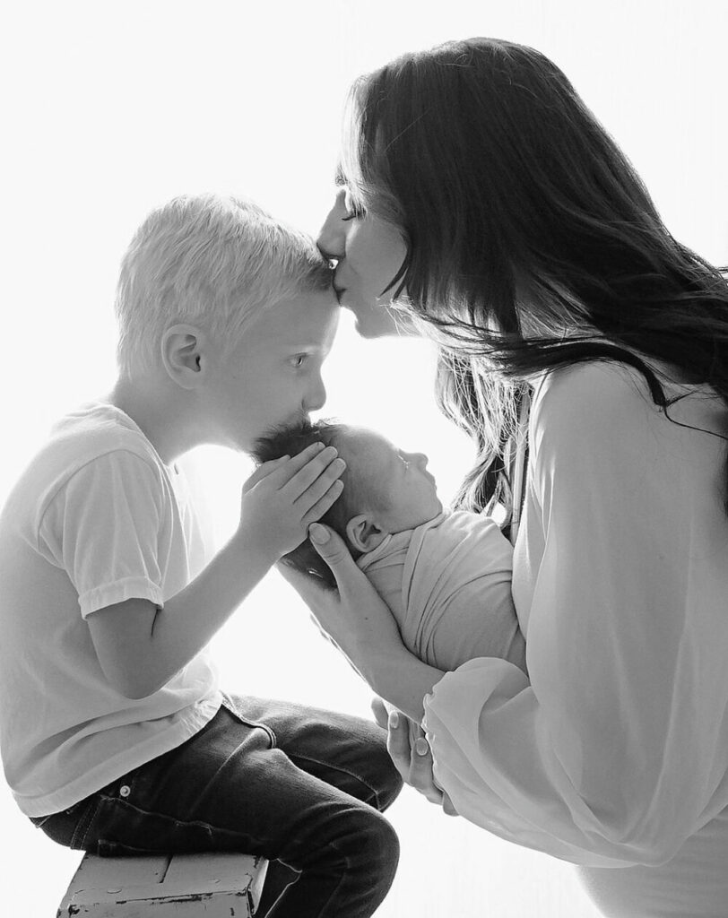 Black-and-white portrait of a mother, kissing her toddler son’s forehead as he kisses his infant brother’s head while they pose for their in studio newborn session in Lumberton, New Jersey.