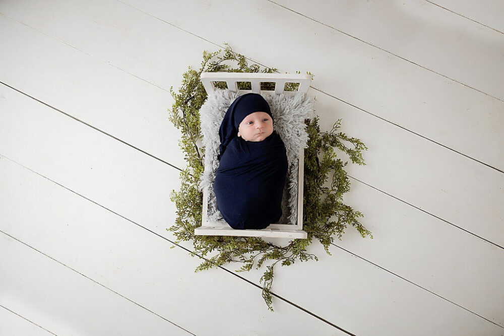 Infant portrait of a boy, wrapped in swaddle, laying on textured blanket in tiny crib, with greenery for their newborn photography near me in Pemberton, New Jersey.