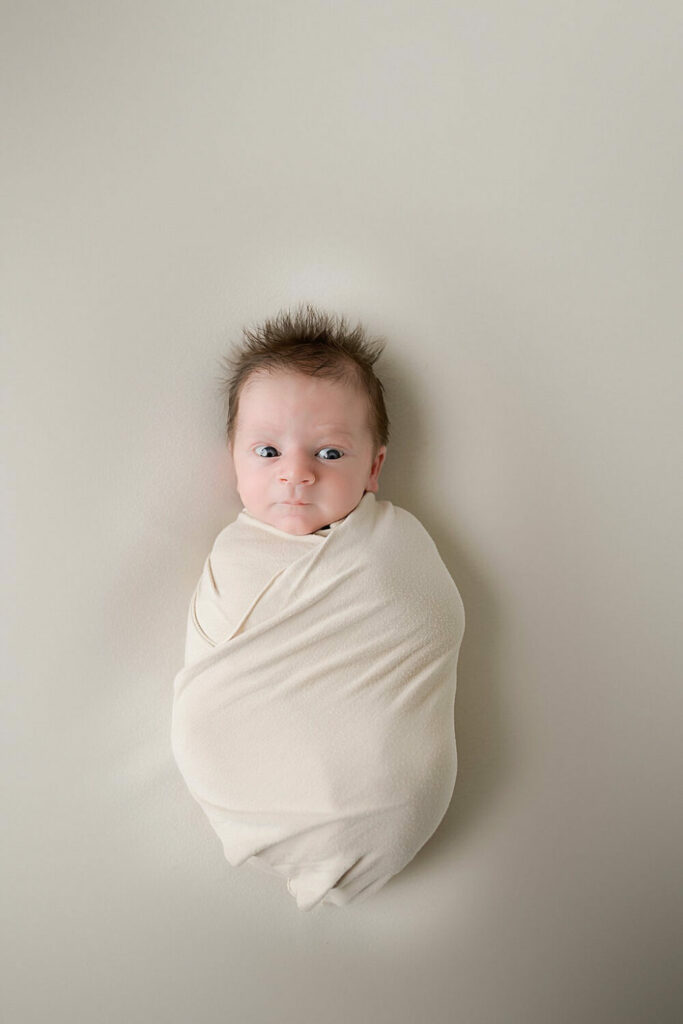 An infant laying on back as he is wrapped in a swaddle awake for his baby photography taken during his newborn session in Medford, New Jersey.