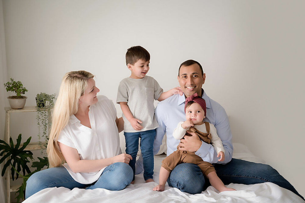 Lifestyle portrait of a man and a woman sitting on the bed photography set with their two kids wearing light colored shirt and denim for their family images taken in Eastampton, New Jersey.