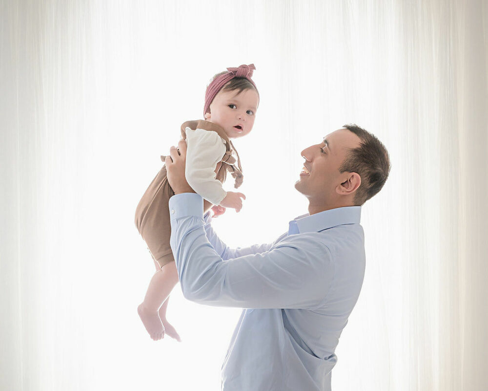 A man looking and holding his daughter in the air against a light and bright background for it their in-studio family session in Westampton, New Jersey.