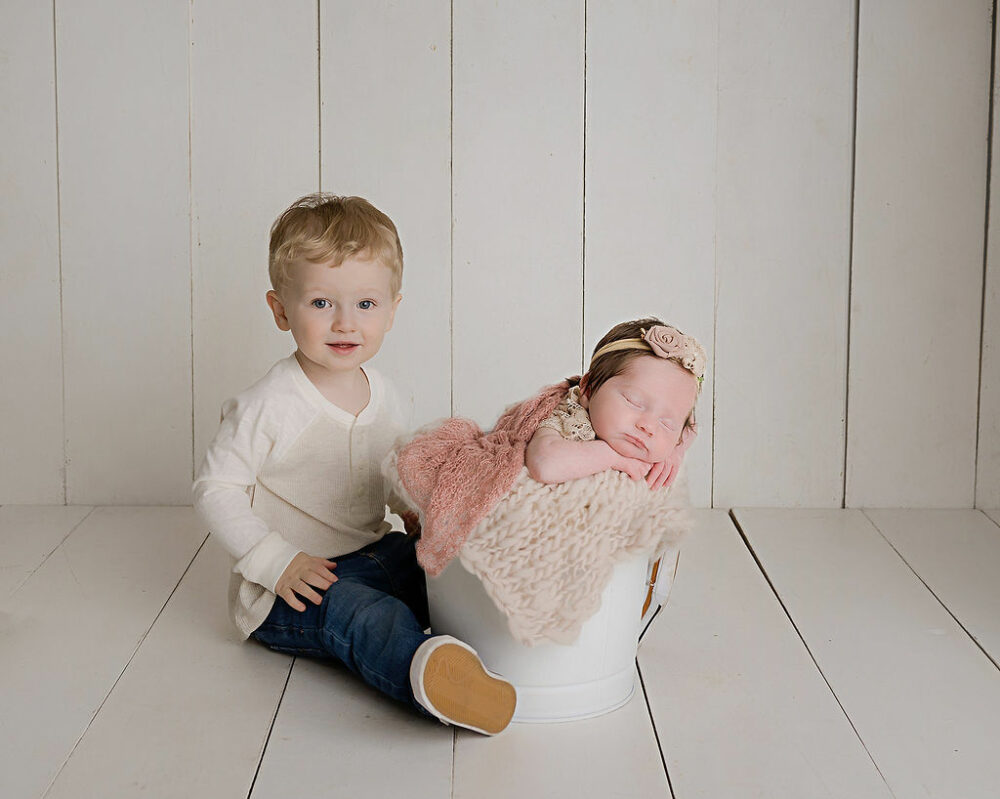 A big brother sitting next to his sleeping, baby sister, Ash, she rest in a photography prop for her airy newborn session in Monroe, New JErsey.