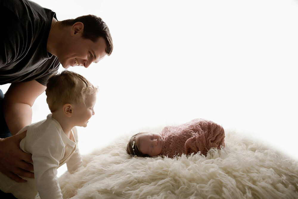 A man, smiling, and holding his toddler son as they watch his baby sister sleep during her in-studio airy newborn session in Bridgeton, New Jersey.