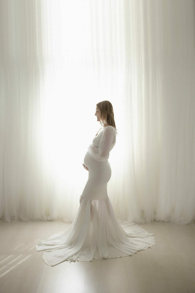 A profile picture of a woman standing, smiling and holding her belly doing photography poses and a long maternity gown for her maternity session in Mount Holly, New Jersey.