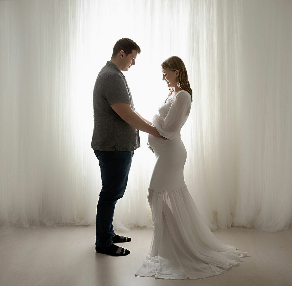 A men and women facing each other as a stand in front of a light and bright background for their couple maternity photos taken during a maternity session in the Vincentown, New Jersey.