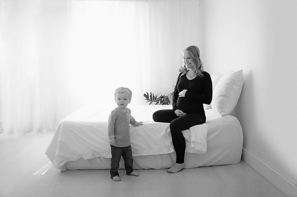 Woman sitting on bed smiling and holding her belly as her son stands on the floor for her black-and-white maternity session in Southampton, New JErsey.