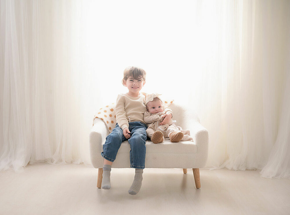 Sibling pictures of a boy and girl sitting on small sofa photography prop against a light and bright backdrop wearing jeans and knit outfits for their portrait photo shoot session in Medford, New Jersey.