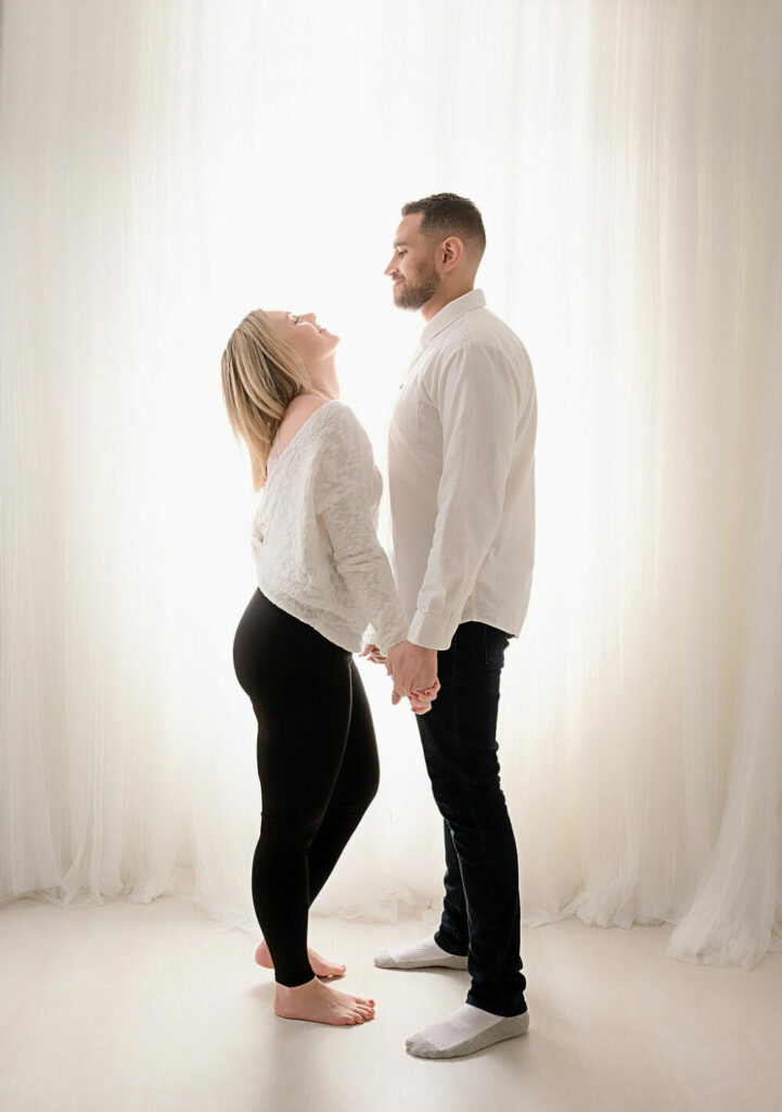 Couple portrait of a man and woman facing each other while they hold hand and looking into each others eyes wearing matching outfits against a light and bright backdrop for their family portrait clothing ideas in Tabernacle, New Jersey.