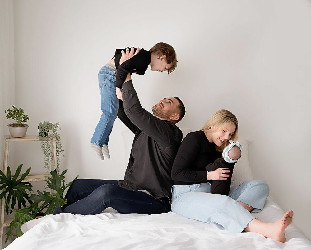 Family photos of a man lifting his toddler in the air while a woman hold her infant for family photo ideas taken during their lifestyle in studio session in Mount Holly, New Jersey.