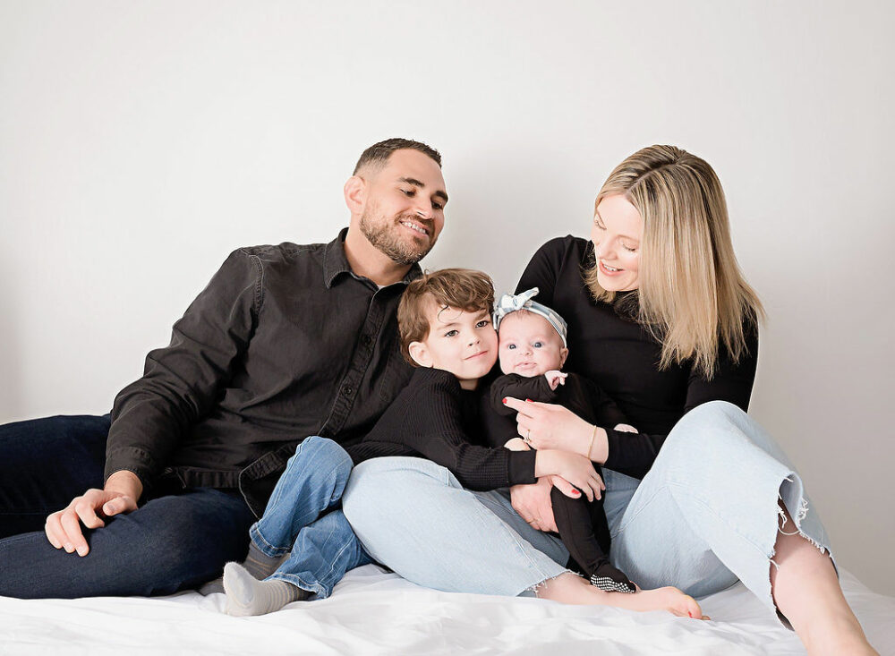 Family portrait of a man and woman sitting on bed photography prop with their two kids wearing black and denim for their lifestyle in-studio session in Lumberton, New Jersey.