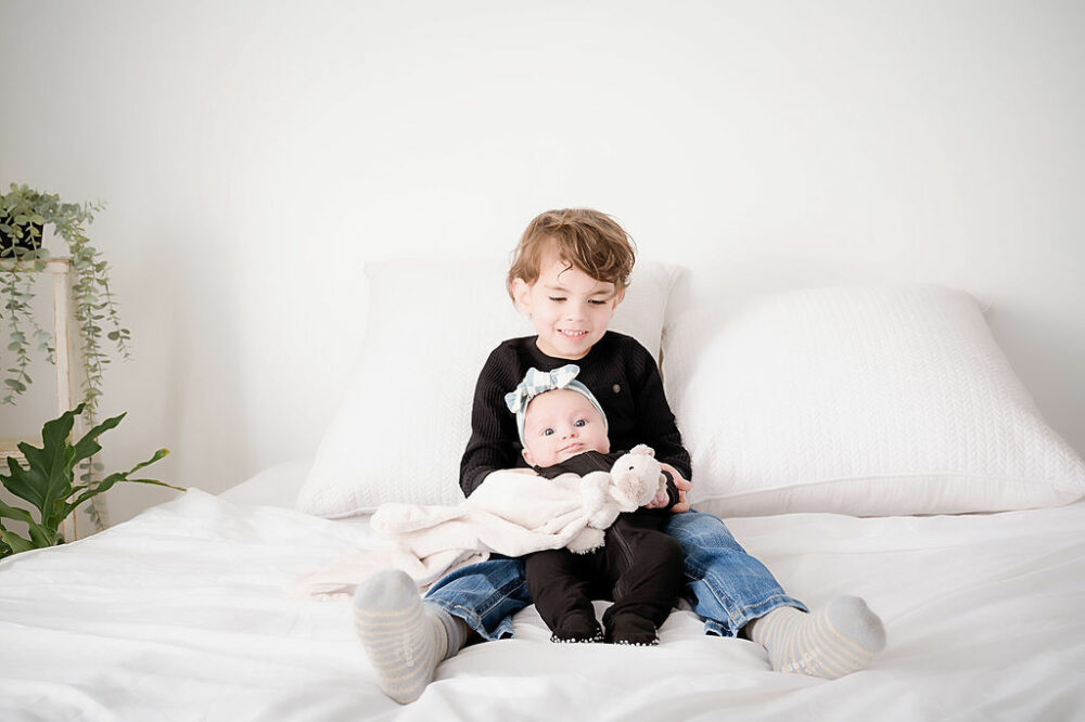 A toddler boy sitting on bed photography prop with his infant sister posing for their sibling pictures taken in Camden, New Jersey.