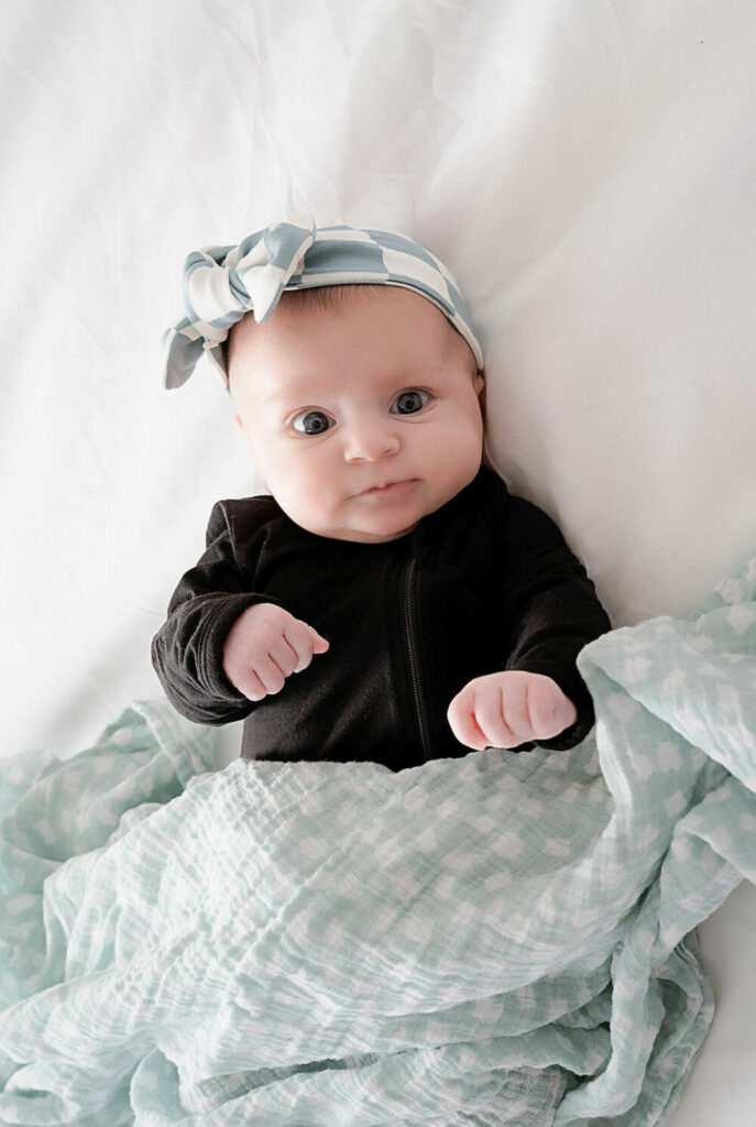 A two month old baby laying on her back wearing long sleeve onesie and covered with a blanket looking at camera for her newborn photography session in Southampton, New Jersey.
