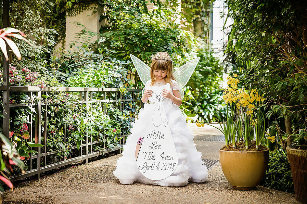 Mile stone photo shoot of a girl, carrying personalize, heirloom, wearing dress and fairy wings standing in garden for her birthday photo shoot in Longwood Gardens, New Jersey.