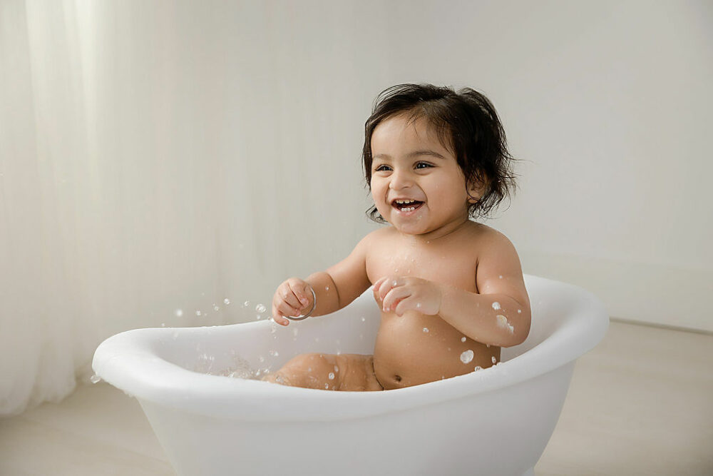 Portrait of a girl, smiling as she sets in bathtub, photography prop against a light backdrop for her first birthday session in Southampton, New Jersey.