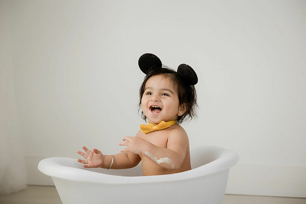 Picture of a toddler wearing bowtie and ears, smiling and playing in bathtub, photography prop for her in studio kick smash session in Eastampton, New Jersey.