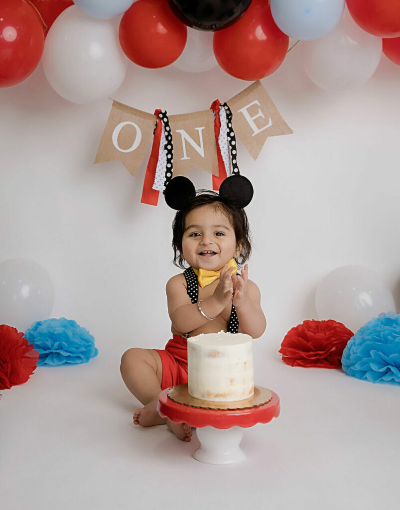 Custom photography set with Mickey Mouse theme adorned with balloons and a one-year-old birthday cake for her Mickey Mouse cake smash session in Hamilton, New Jersey.