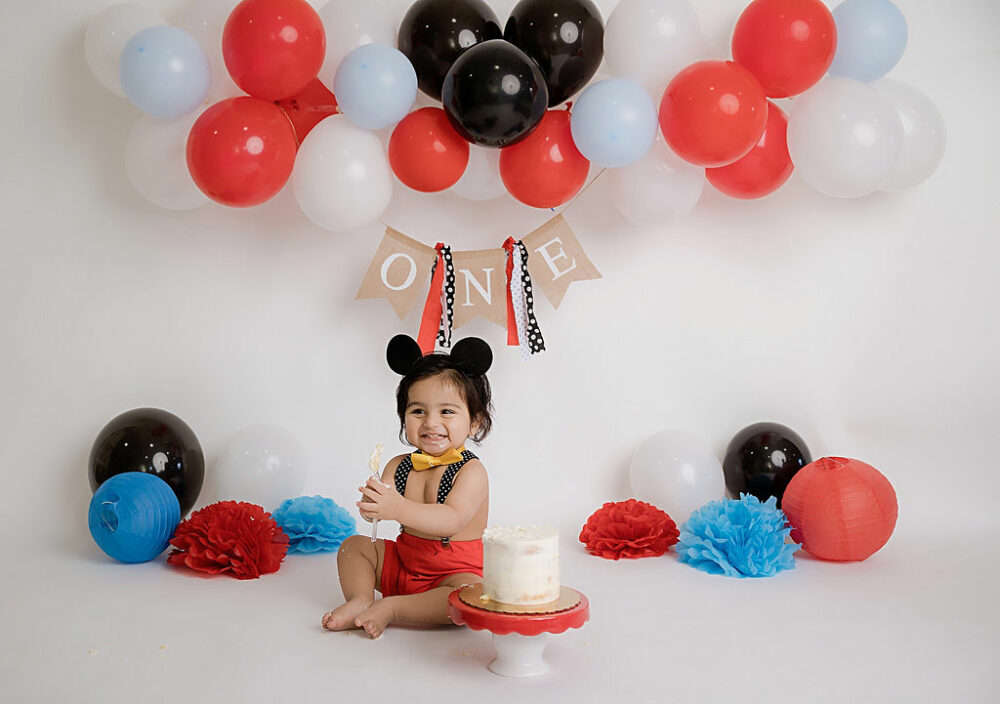 Milestone photography of a one-year-old girl, smiling and sitting behind a cake for her Mickey Mouse cake smash session in Pemberton, New Jersey.