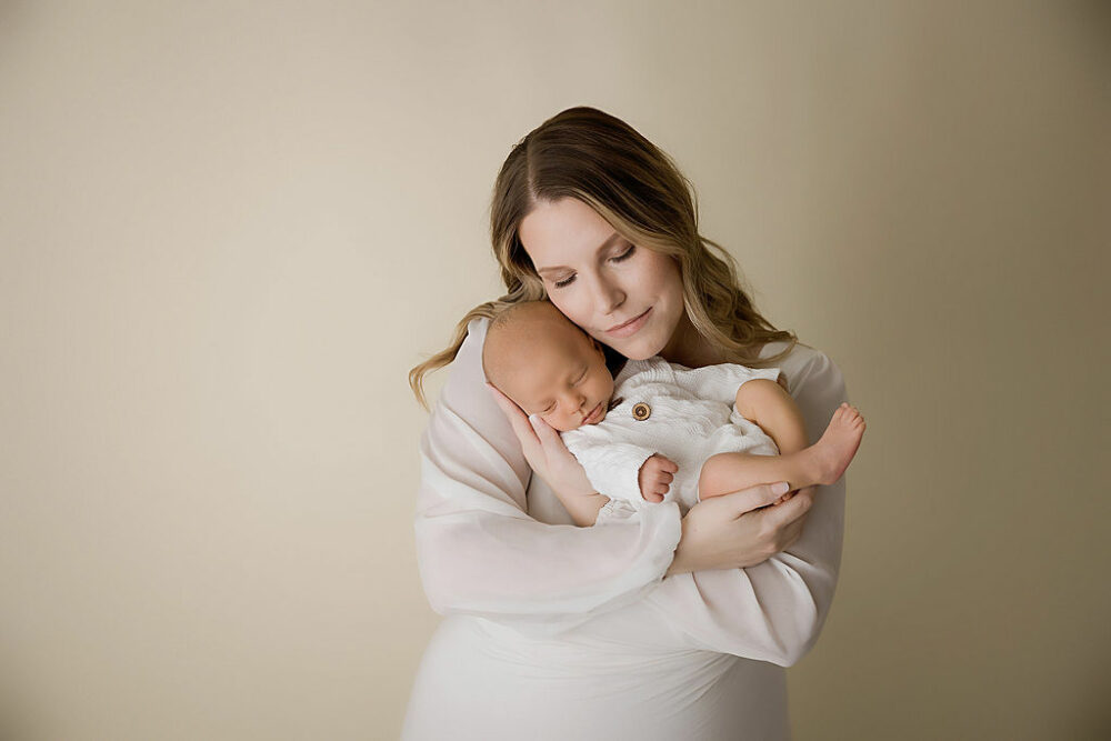 A woman standing and holding her infant boy posing for their infant photography session with a cream backdrop, and wearing white clothes for their in-studio newborn session in Camden, New Jersey.