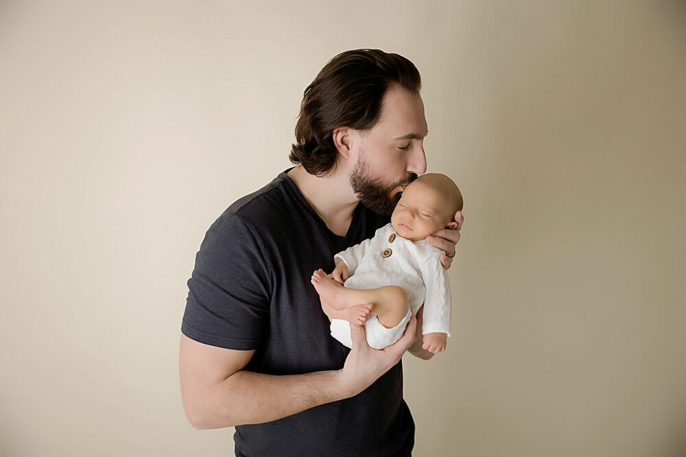 A man holding his infant against his chest and kissing his head for a father and son infant photos, taken during their in-studio, navy blue newborn session in Mount Holly, New Jersey