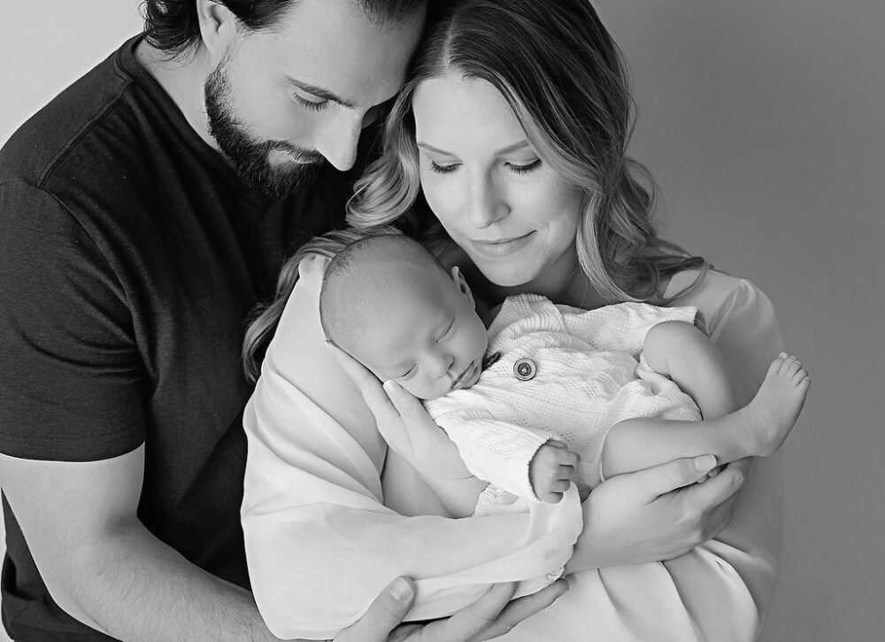Black and white family portrait of a man and woman, hugging and holding their newborn son as he sleeps for their in studio, navy blue newborn session in Hamilton, New Jersey.