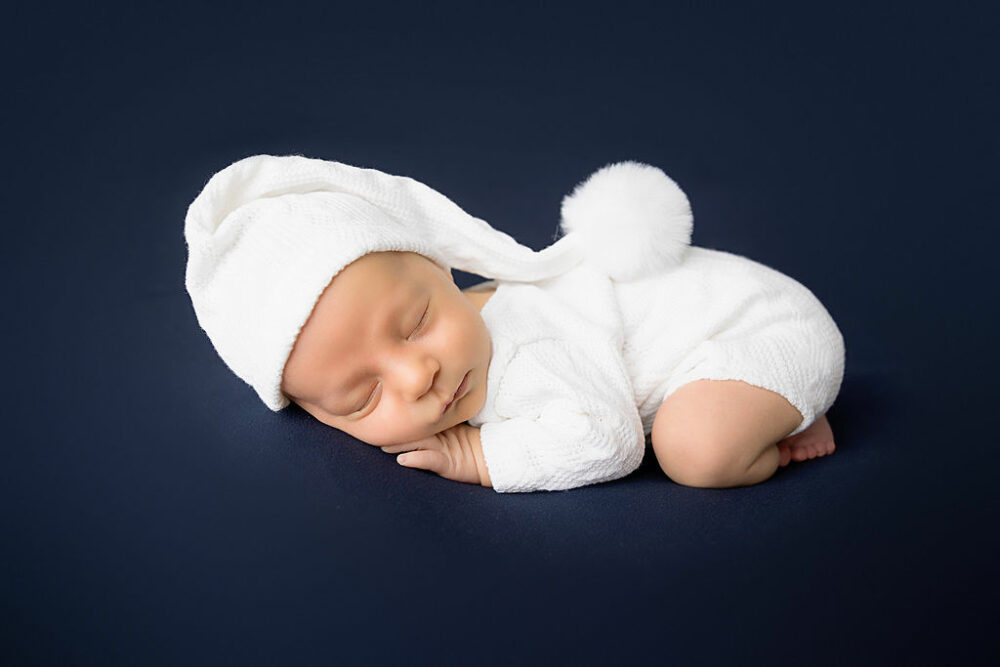 A sleeping boy, dressed in matching outfit and hat laying on tummy for a in-studio navy blue newborn session in Eastampton, New Jersey.